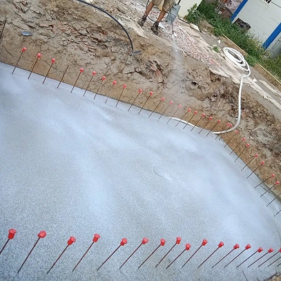 Application de béton projeté (shotcrete) dans une excavation, des tiges métalliques avec capuchons rouges marquent l'épaisseur de la future piscine ou structure.