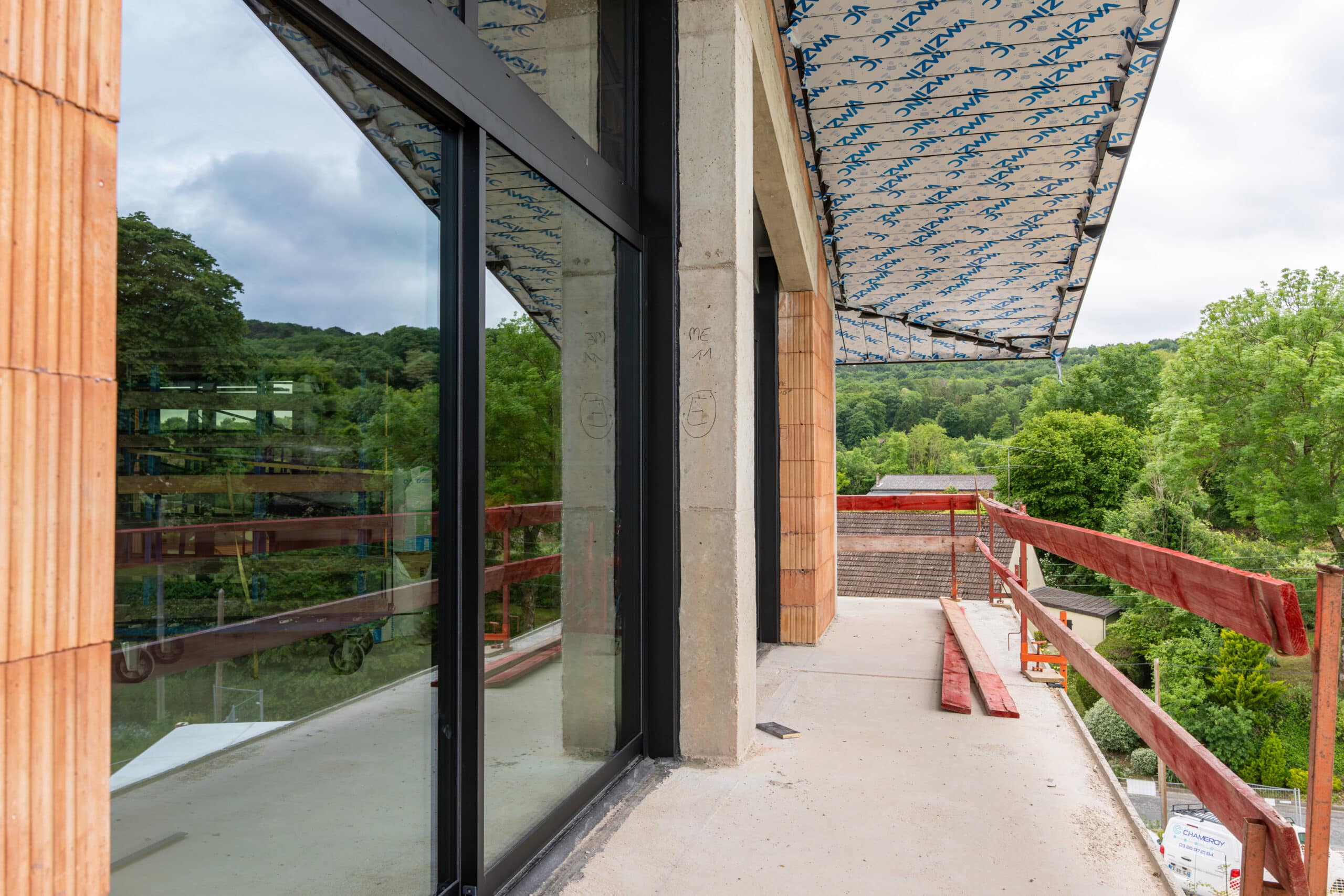 Chantier de construction moderne : grande baie vitrée noire, pilier en béton, mur en briques terre cuite, et sous-face avec isolation 'ZWAZINC'.