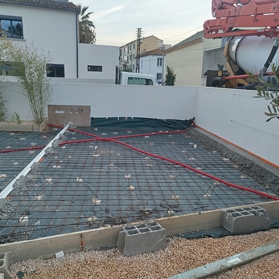 Préparation de dalle de béton avec treillis soudé, tuyaux rouges (chauffage au sol ?), coffrage et camion pompe en arrière-plan.