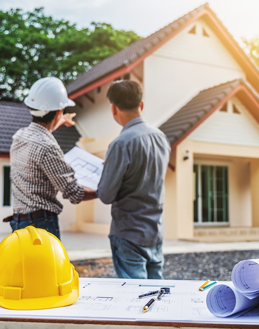 Architecte et client discutent plans devant maison neuve, casque jaune et plans sur table au premier plan.