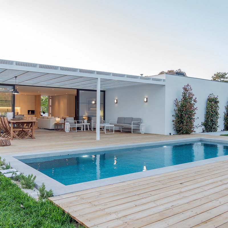 Moderne maison avec terrasse en bois clair, pergola blanche, salon extérieur, et piscine rectangulaire aux eaux bleues, éclairée au crépuscule.