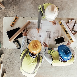Trois ouvriers en gilets et casques (blanc, jaune, bleu) examinant des plans d'architecture sur une table, pointant du doigt des détails techniques.