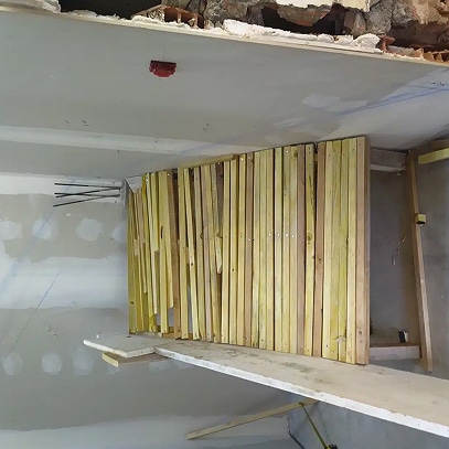 Structure de bois clair en construction, peut-être pour une cloison ou un revêtement mural, avec un plafond en plâtre brut et une boîte électrique rouge visible.