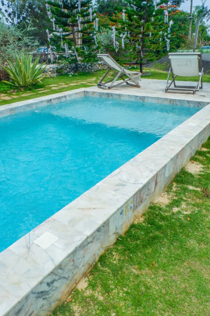 Piscine en Marbre et Jardin Tropical Relaxant Piscine rectangulaire aux margelles en marbre blanc, entourée de gazon vert et de végétation luxuriante avec des chaises longues en arrière-plan.