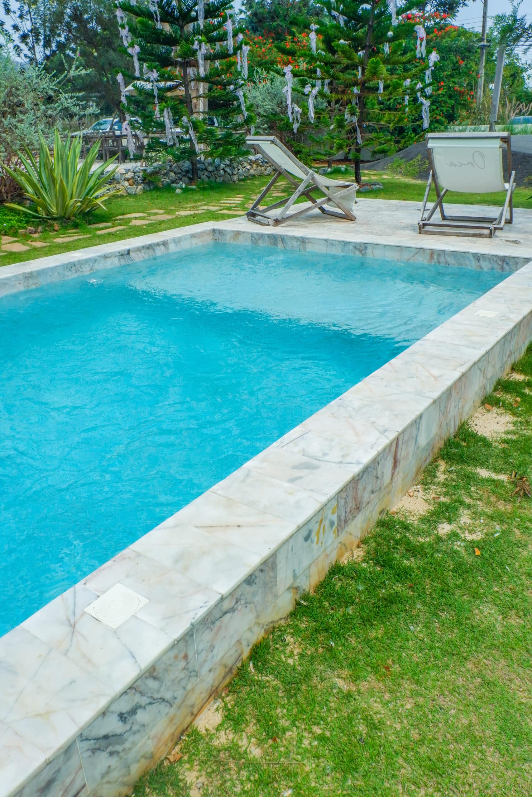 Piscine rectangulaire aux margelles en marbre blanc, entourée de gazon vert et de végétation luxuriante avec des chaises longues en arrière-plan.