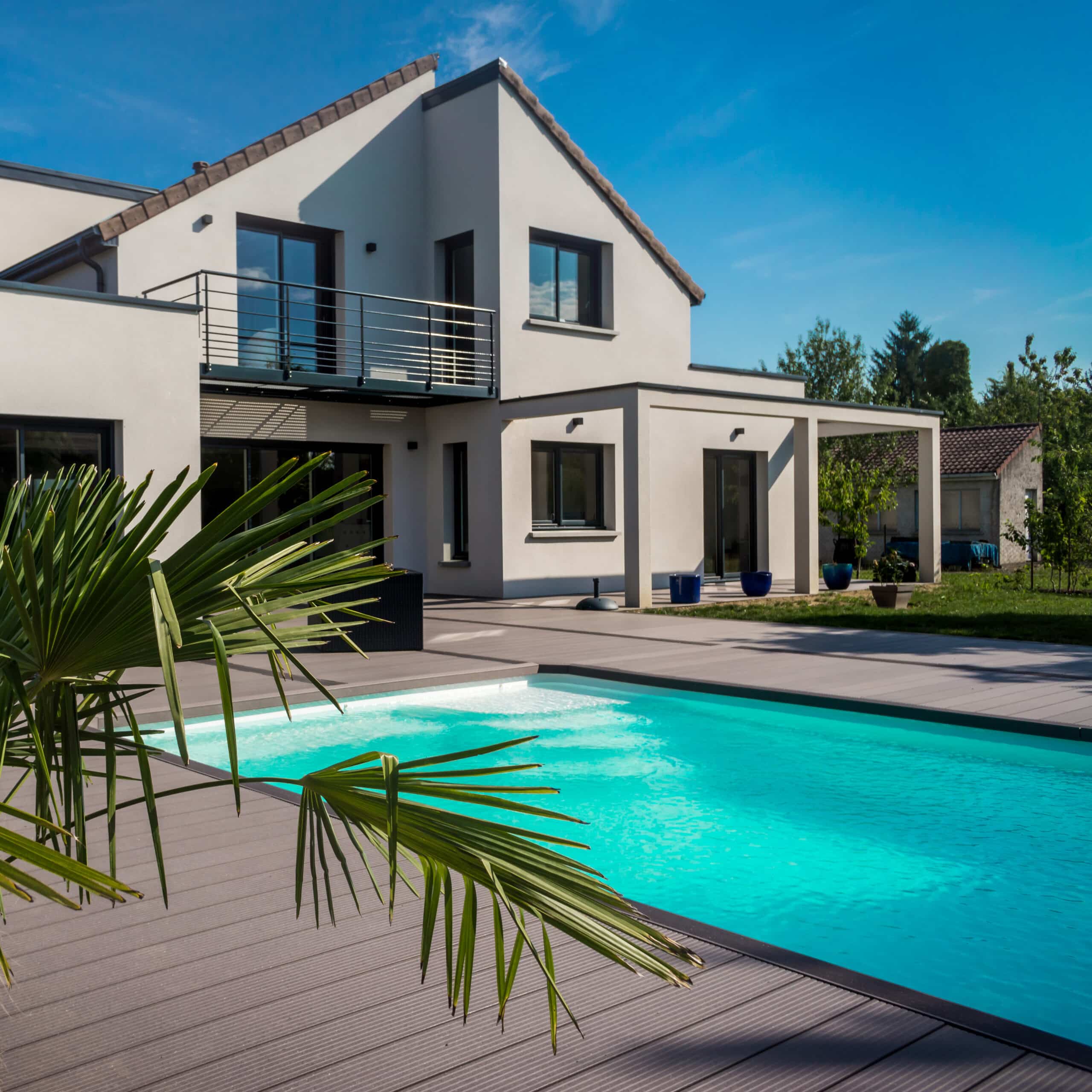 Piscine moderne avec terrasse en bois composite, devant une villa contemporaine blanche sous un ciel bleu vif, palme au premier plan.